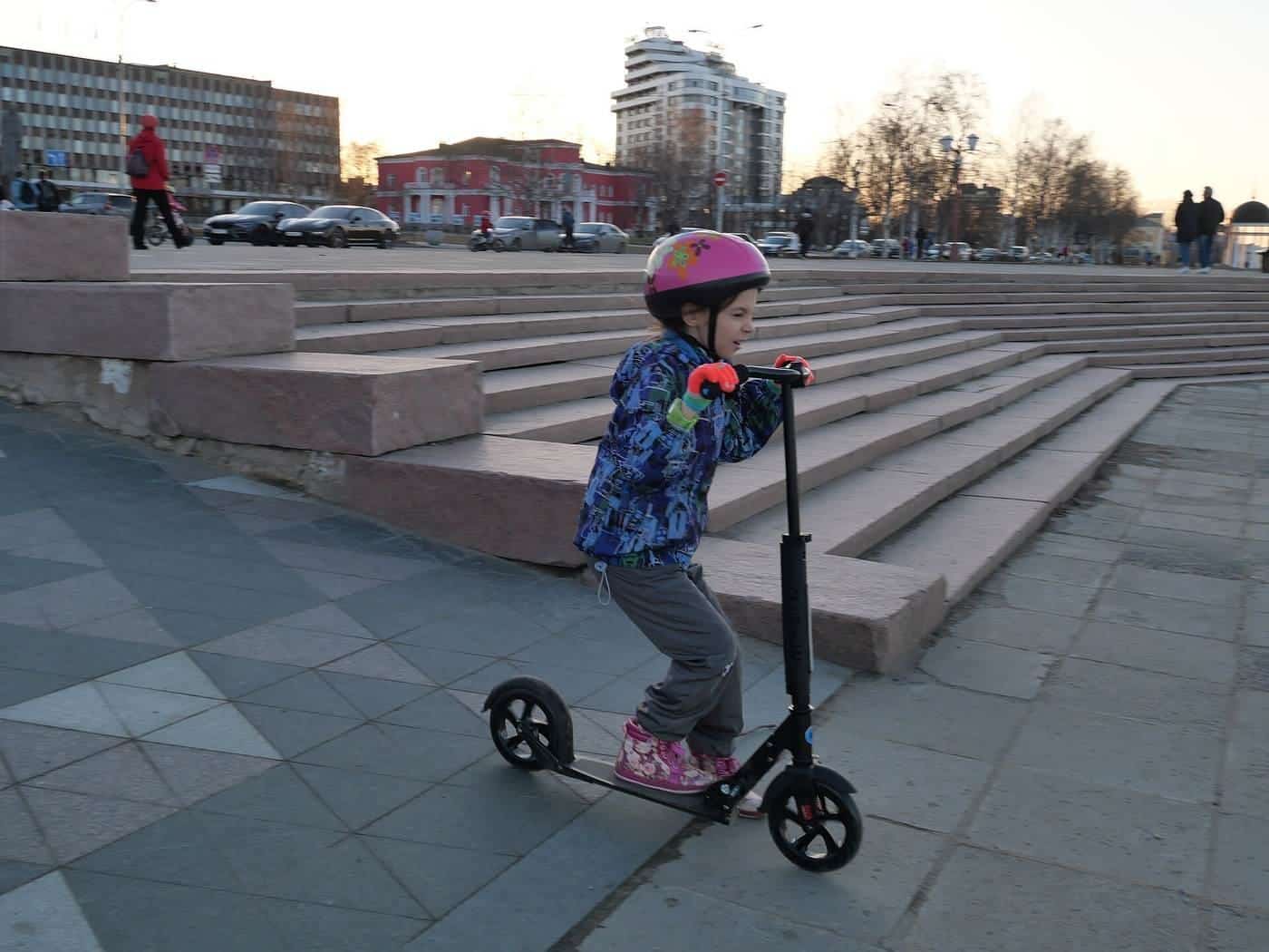 A young child in a pink helmet riding a kick scooter on a paved plaza
